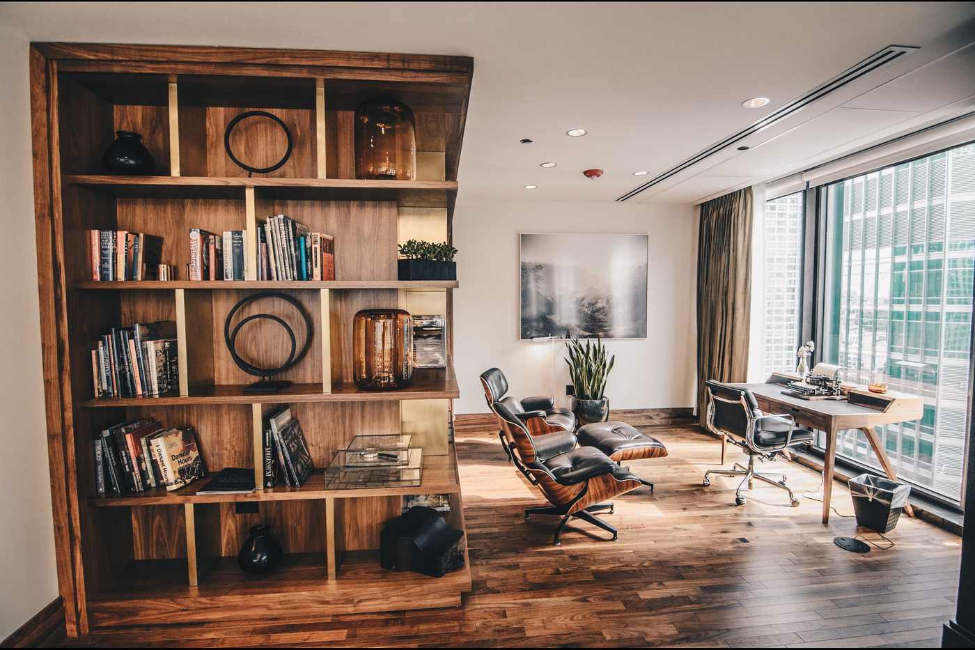 A private office after hours: a walnut bookshelf, a desk by the window and a lounge chair, with nobody in the room.